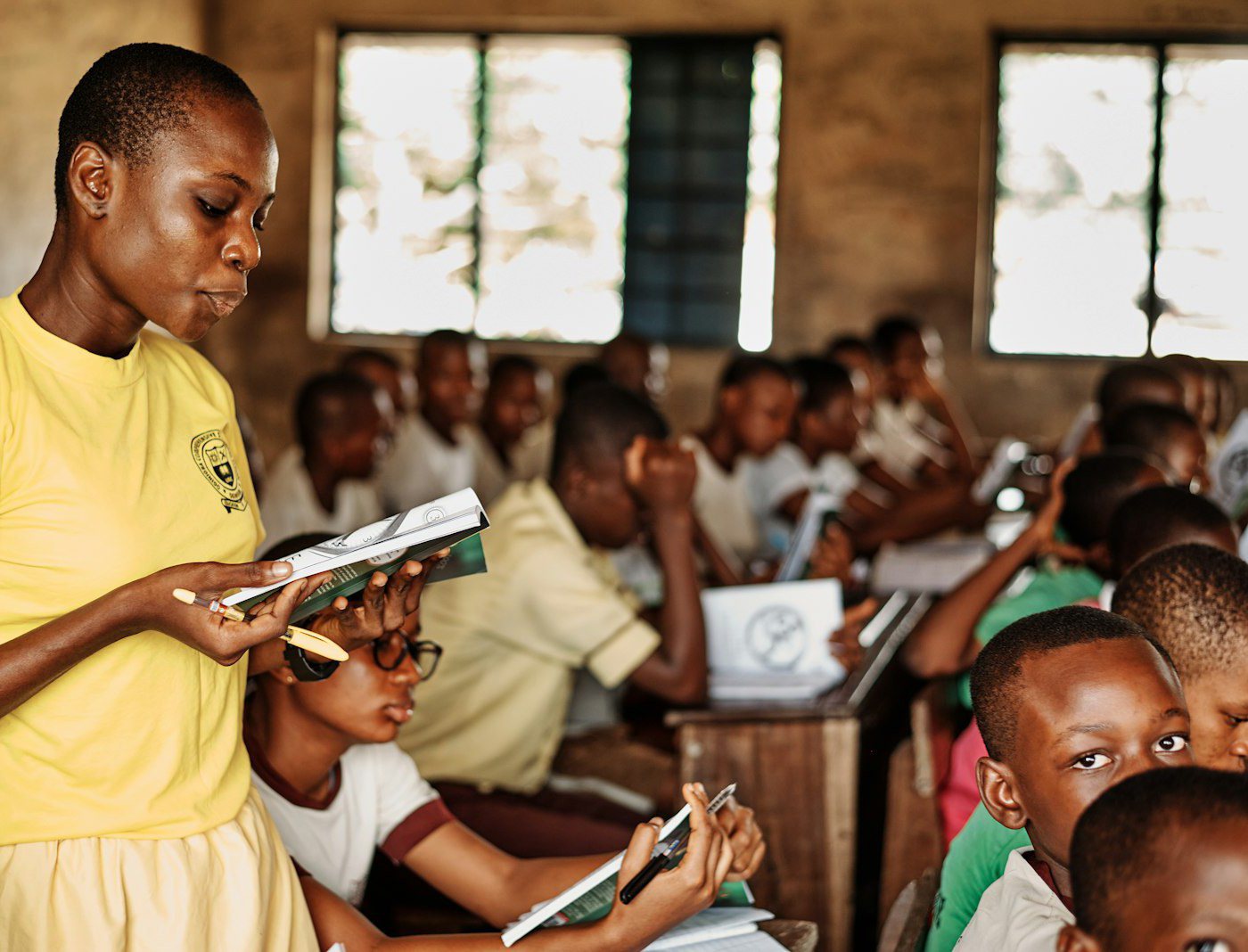 A teacher and pupils in a Nigerian classroom, working from exercise books and a paper register